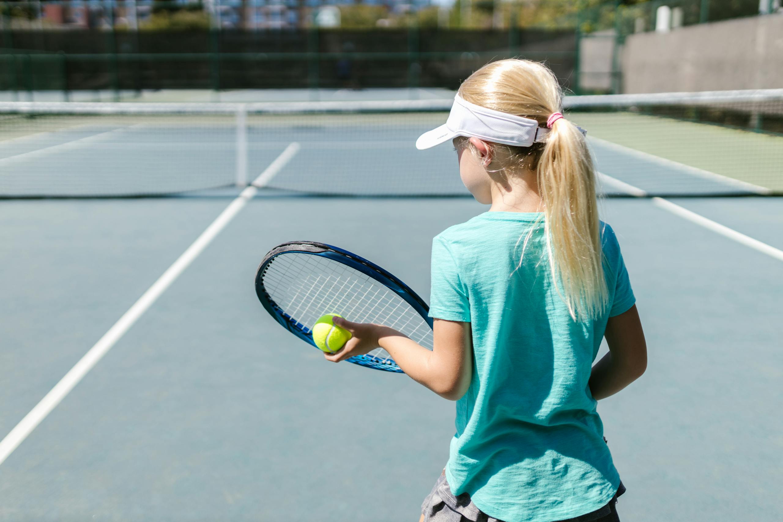 A young girl holding a tennis racket and ball on an outdoor court in daylight.